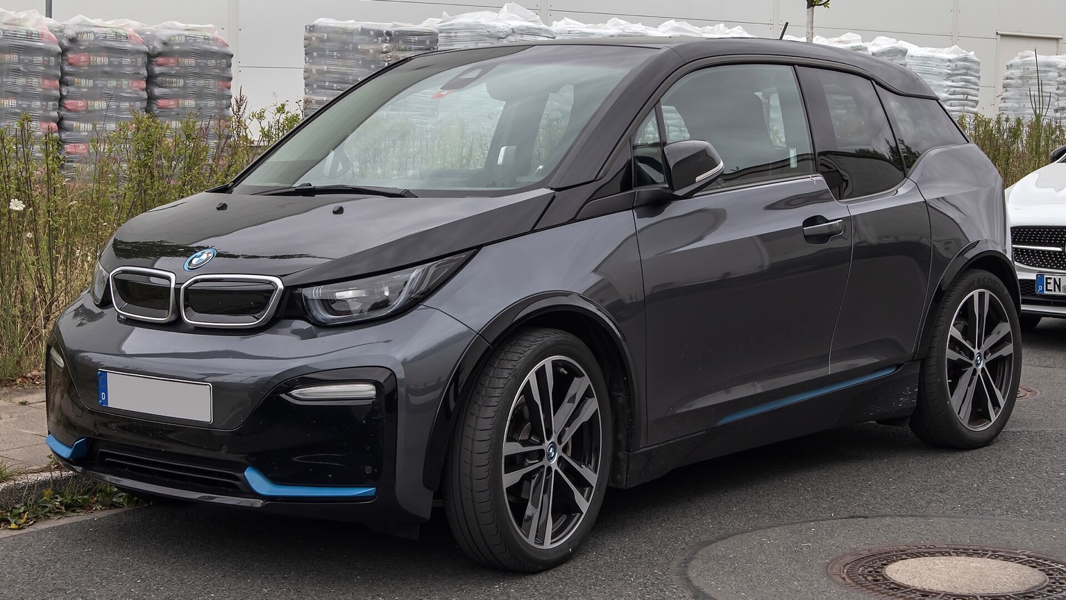 Szürke, modellfrissítés utáni BMW i3 elektromos autó elölről és oldalról, parkolóhelyen fényképezve